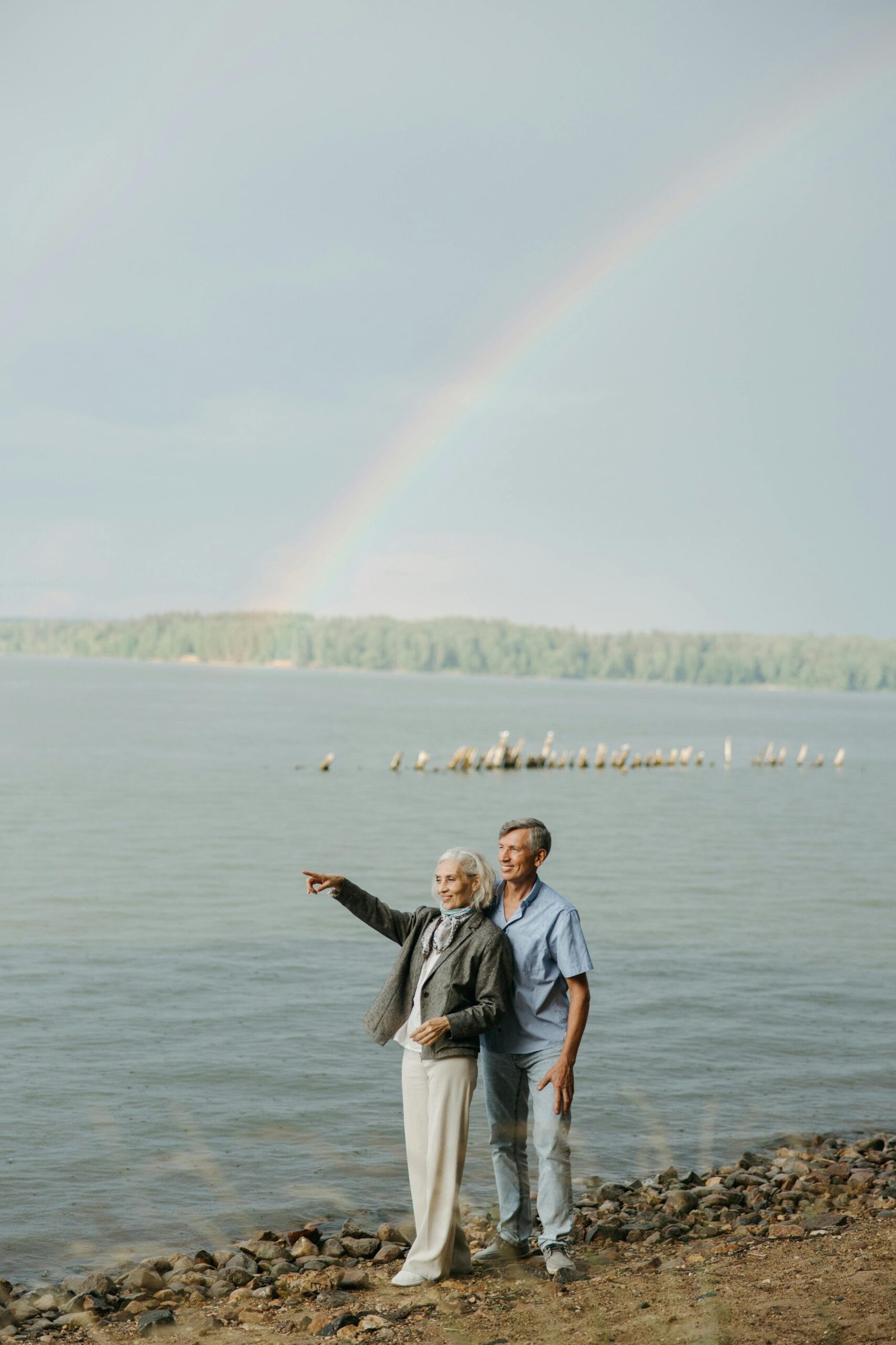 Elderly couple embraces by the lake under a vibrant rainbow, enjoying nature together.